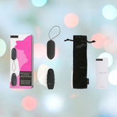 A pink and black box features the silhouette of a woman's profile next to the words "B Swish Bnaughty Classic Unleashed - Black." Nearby, there's a black remote-controlled bullet vibrator, a sleek drawstring pouch, and an informative instruction booklet. The blurred background shimmers with colorful bokeh lights.
